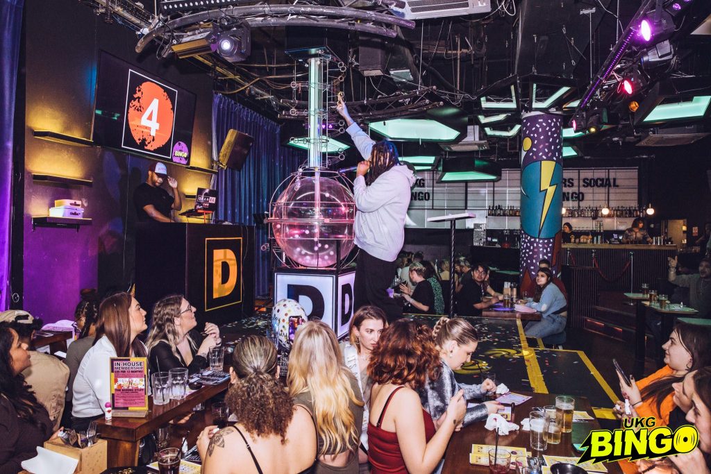 Bingo crowd enjoying the game UKG Bingo event atmosphere Excited participants marking bingo cards Colorful bingo card close-up Grand prize announcement moment Voucher prize showcase Flight ticket prize display Music vinyl prize collection Unique party gifts for winners UKG Bingo logo and branding Happy winner celebrating victory Event host or DJ entertaining the crowd High-energy crowd shot at UKG Bingo Diverse audience engagement in bingo Bingo cards arranged for gameplay